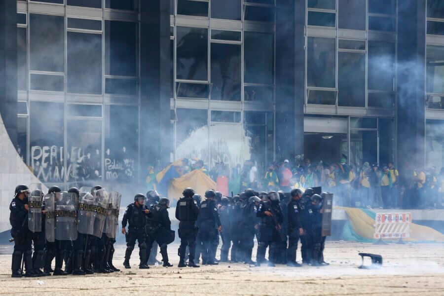 Policiais em primeiro plano; ao fundo, golpistas depredando prédios públicos no 8 de Janeiro
